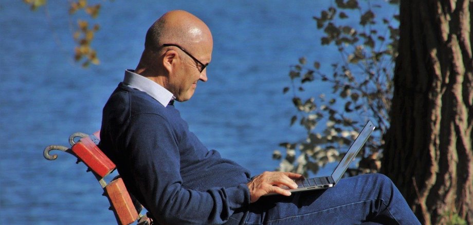 Ein älterer Mann mit Glatze sitzt an einem See und surft mit einem Laptop im Internet