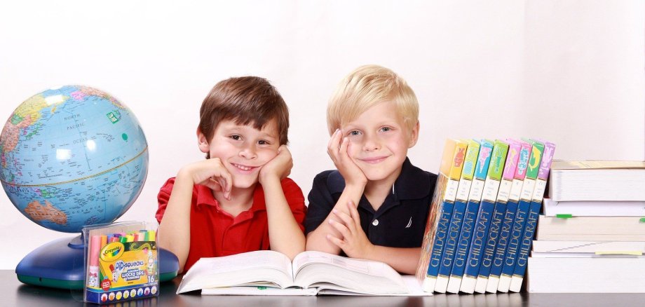 Zwei Jungs an einem Schultisch. Vor ihnen Bücher und ein Globus. Die Kinder halten Stifte und lachen.