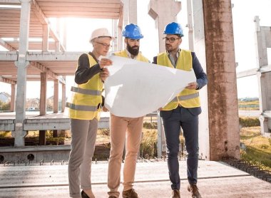 Das Foto zeigt symbolisch zwei Männer und eine Frau auf einer Baustelle. Sie tragen Bauarbeiterhelme und beugen sich gemeinsam über einen großen Bauplan. 
