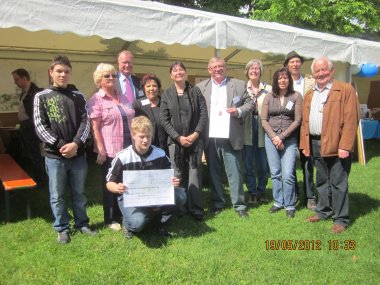 Eine Gruppe lachender Menschen steht auf einem Rasen. Vorn links kniet ein Jugendlicher mit einer Urkunde in den Händen.