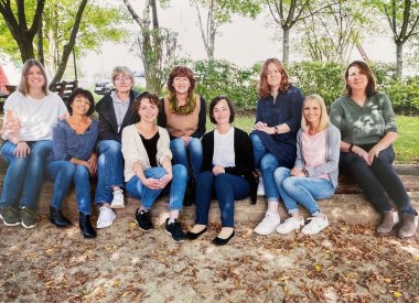 9 Erzieherinnen sitzen im Freien auf einem Baumstamm. Sie sind das Team der Kita Roth. Alle lächeln. Im Hintergrund sind Bäume zu sehen.